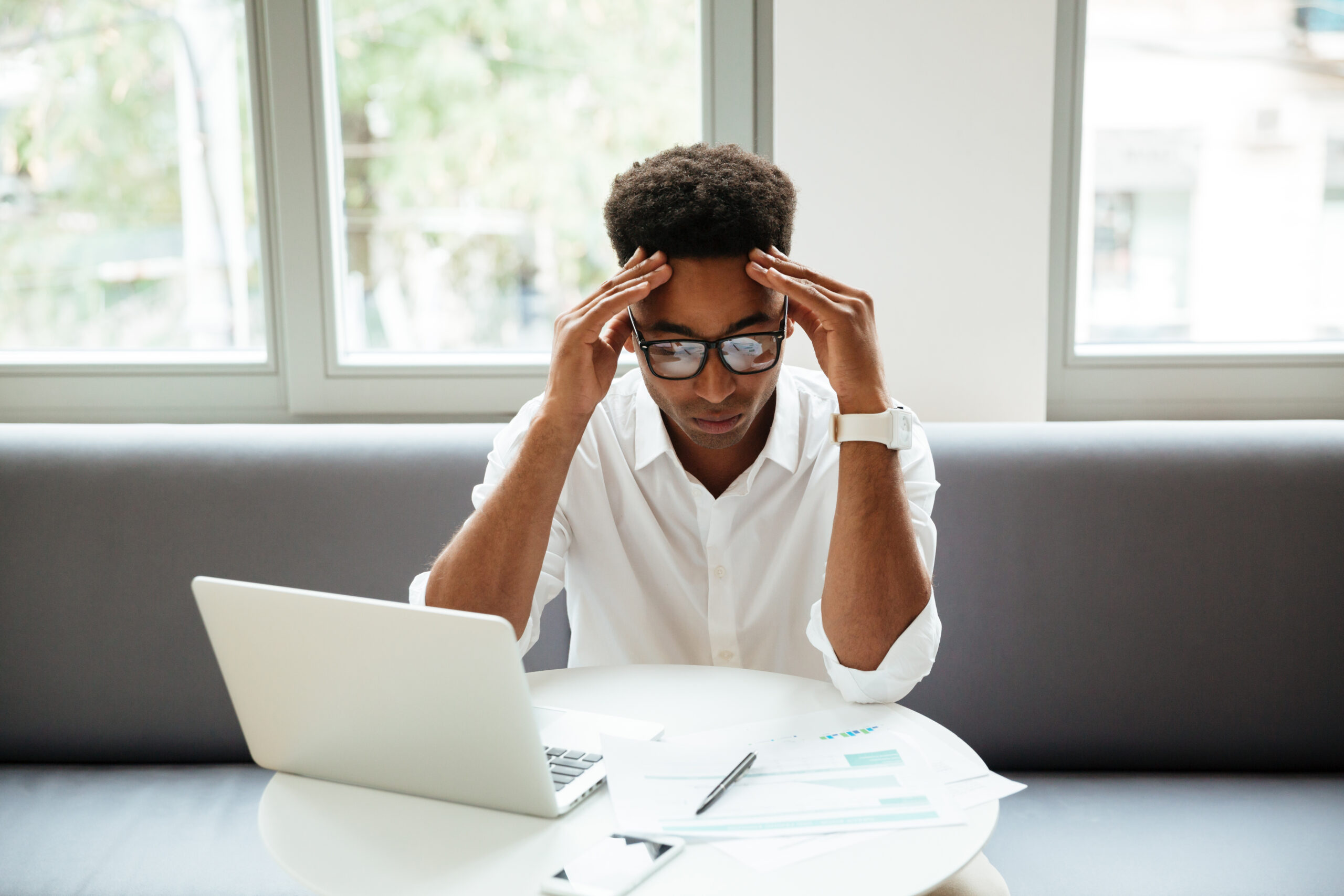 Picture of concentrated serious young african man sitting coworking by laptop computer near documents. Looking aside.