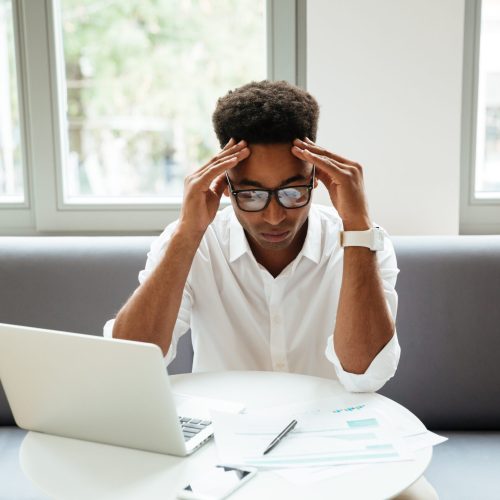 Picture of concentrated serious young african man sitting coworking by laptop computer near documents. Looking aside.